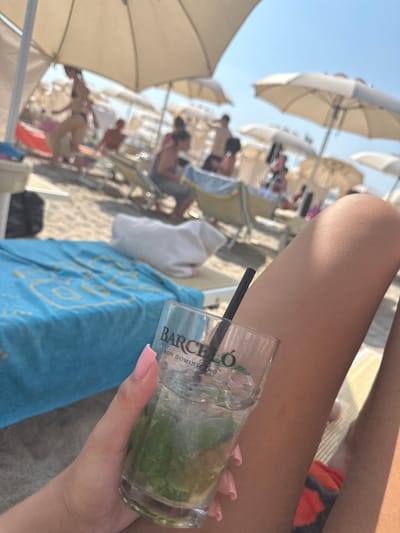 A first-person perspective shot shows a woman's hand holding a glass of iced drink with mint and lime on a sunny beach. In the background, people relax under umbrellas on lounge chairs. The scene is set in Finale Ligure, Italy, a popular coastal town known for its beaches and clear waters. The woman's legs and part of a bikini are visible in the foreground, suggesting a leisurely day at the beach. The overall atmosphere is relaxed and summery.
