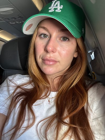 A woman with long, auburn hair and fair skin is seated on an airplane. She is wearing a bright green baseball cap with a white "LA" logo and a white t-shirt. The cap is angled to partially obscure her forehead and eyes, but her right eye, with a greenish-blue iris, is visible. Her skin appears slightly flushed and reflective, possibly due to airplane cabin lighting or makeup. She has a small dimple visible on her right cheek. She is wearing a delicate necklace with a small pendant. The background shows the interior of an airplane cabin, with a dark gray airplane seat to her left and the overhead compartment and air vents visible above. The lighting is soft and appears to be from an artificial source, typical of an airplane cabin. The image is a close-up selfie, with the woman's face and upper chest filling the frame. The location is not determinable from the image.