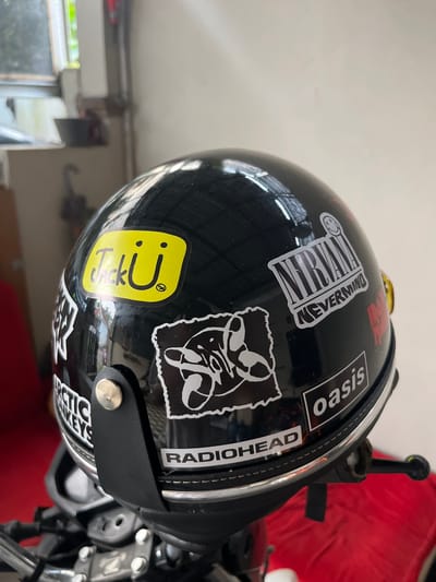 A black, glossy motorcycle helmet adorned with various stickers is the central subject of this image, captured in Paweden, Indonesia. The helmet is positioned on what appears to be a motorcycle or scooter, with parts of the handlebars and metallic components visible at the bottom right.

Several band stickers are prominently displayed on the helmet. A rectangular, black and white sticker for "NIRVANA" with its iconic smiley face logo and "NEVERMIND" text is visible on the top left. To its right, a white rectangular sticker with black text reads "oasis". Further right, along the edge of the helmet, another white text sticker vertically spells out "RADIOHEAD" on a black background. A distinct yellow sticker with rounded corners features black text "TikU" and a small circular logo above the 'U'. Additionally, a large black and white sticker depicts an abstract, somewhat anthropomorphic figure, contributing to the helmet's personalized appearance.

The helmet's reflective surface shows glimpses of its surroundings, including a light-colored ceiling or wall above and a window with some outdoor greenery in the lower left background. A red object, possibly a part of the motorcycle's seat or a cover, is visible in the upper right background. The lighting suggests an indoor setting during daylight hours. The image focuses on the helmet and its unique decorations, with no human subjects or explicit activity taking place.