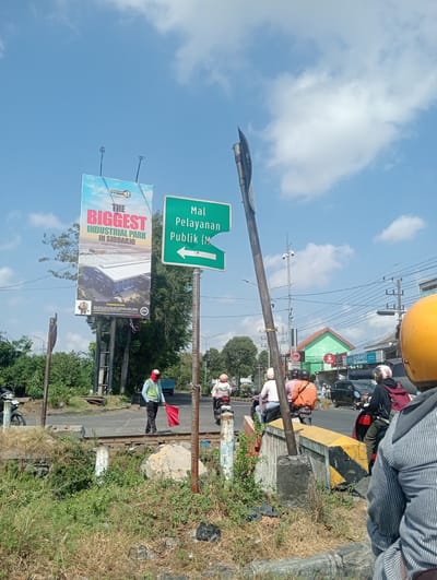 The image depicts a street scene in Banjarkemantren, Indonesia, with a blue sky and scattered clouds. A large billboard advertises "The Biggest Industrial Park in Sidoarjo." To the right, a green directional sign points left, indicating "Mal Pelayanan Publik (M)." A traffic controller in a high-visibility vest and red head covering stands on train tracks, holding a red flag. Several motorcycles and their riders are visible on the road, suggesting traffic. Vegetation lines the sides of the road, and some construction or infrastructure elements, including yellow and black striped barriers, are present. The overall impression is of a busy intersection in an urban or semi-urban setting.