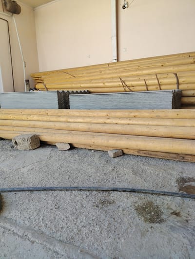 In Almachi, Uzbekistan, a storage area is depicted, with a concrete floor and pale beige walls. Stacked horizontally are numerous large, light-colored wooden logs, some appearing unfinished. In front of the logs, several sheets of corrugated roofing material are neatly stacked. A dark electrical cable lies across the concrete floor. The environment appears to be indoors, possibly a warehouse or a covered outdoor space, with an overhead light fixture and an electrical outlet visible on the wall.