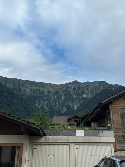 In Bönigen, Switzerland, the image captures a scene dominated by a majestic mountain range. The foreground features the roofs and facades of several buildings, including a house with a wooden exterior and a dark-tiled roof, and a structure with a light-colored garage door. The mountain, covered in dense green forest and patches of rocky terrain, looms in the background. The sky above is a mix of white clouds and blue hues. A dark SUV is partially visible on the right side of the frame. The overall impression is of a serene, natural landscape juxtaposed with human habitation.