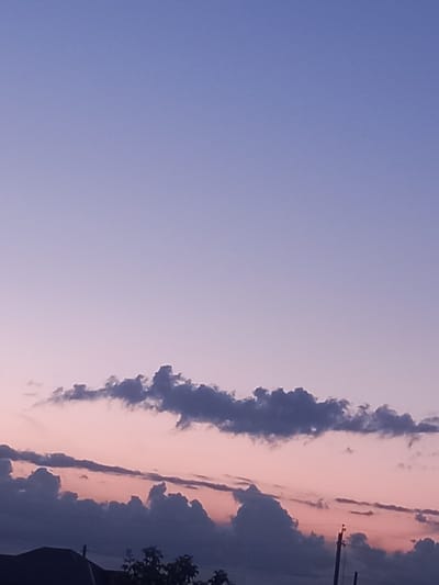 The image depicts a twilight sky over a rural landscape in Қозыкеткен ауылдық округі, Kazakhstan. The sky transitions from a light purple hue at the top to softer pinks and purples near the horizon, indicating either sunrise or sunset. Several dark, cloud formations are visible against the colorful sky. In the foreground, the silhouette of a building roof and some trees can be seen, suggesting a residential or agricultural area. A utility pole with wires is also present, further confirming a populated or developed area. The overall scene is tranquil and atmospheric, characteristic of a rural evening or early morning.