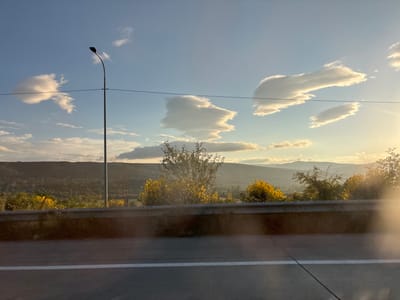 This is a landscape photograph taken from a moving vehicle, likely on a highway. The scene depicts a rural setting in Changilari, Georgia, during what appears to be late afternoon or early evening, judging by the warm, golden light and long shadows. The sky is a clear blue, dotted with distinct, sculpted clouds that are illuminated by the setting sun. A tall, slender lamppost stands prominently on the left side of the frame, its dark silhouette stark against the bright sky. A power line stretches horizontally across the image, bisecting the sky and crossing behind the lamppost.
Below the sky, rolling hills form the background, covered in a dense, green forest. The mid-ground features a vibrant display of yellow-blooming bushes and trees, adding splashes of color to the landscape. A concrete barrier runs along the edge of the road, separating the viewer from the rolling terrain. The foreground consists of a grey asphalt road with a clear white line marking a lane. The overall impression is one of serene natural beauty, with the man-made elements of the road and lamppost integrating into the expansive, sun-drenched vista. There is no visible text in the image.