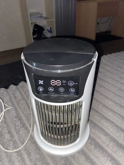 The image shows a white and black portable electric fan with a digital display. The display shows the number "88" and icons for a fan, a light bulb, an atomizer, a timer, and speed settings. The fan is positioned on a gray, textured surface, possibly a carpet or blanket. A white power cord is visible to the left of the fan. In the background, there is a blurred view of shelves with some items on them, and a cardboard box. The scene appears to be indoors. The image does not provide any specific information that would allow for identification of its location in Fălești, Moldova.
