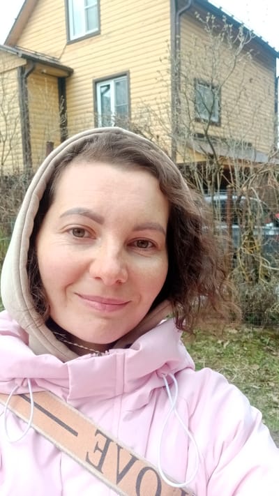 The image is a close-up outdoor selfie of a woman in Былина Снт, Russia. The subject is a woman, likely in her 30s or 40s, with curly brown hair and brown eyes. She is looking directly at the camera with a gentle, pleasant smile.
She is dressed for cool weather, wearing a light pink hooded puffer jacket over a beige hooded sweatshirt, with the beige hood pulled up. A striped fabric strap, possibly from a shoulder bag or purse, diagonally crosses her chest. On this strap, the upside-down letters "OVE" are visible, suggesting part of a word like "LOVE" or a brand name.
The background shows a two-story house constructed with light yellow horizontal wooden siding, dark window frames, and a dark roof. Bare tree branches are prominent around the house and in the mid-ground, indicating either late autumn or early spring. The lighting is soft and even, consistent with an overcast day. The setting appears to be a suburban or rural residential area.