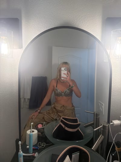 A reflection in a bathroom mirror shows a blonde woman holding a smartphone. The woman is wearing a silver sequined bra top, khaki pants, and jewelry. The bathroom has a white door, a gray towel, a countertop with a sink, and various toiletries including toothbrushes and toilet paper. The location is Palm Desert, USA.
