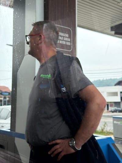 A medium shot shows a man wearing a gray polo shirt with green and blue logos and black pants. He is wearing red-rimmed glasses and a watch on his left wrist. He is standing next to a gas pump, reflected in a window. The background shows a building and trees. The image appears to be taken in Podlugovi, Bosnia and Herzegovina, at a gas station.