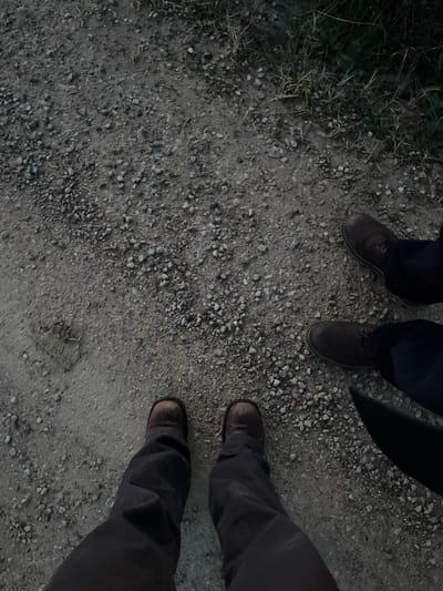 Three individuals are depicted from a high-angle perspective, showing only their lower legs and feet on a gravel path. To the left, one person stands with dark blue trousers and brown lace-up shoes. To the right, two more individuals are positioned; one wears dark grey or brown trousers and sturdy brown boots, while the other's legs and feet are partially visible, also in similar attire.
The scene takes place outdoors on a path composed of small grey and brown gravel mixed with dirt. Sparse patches of green and dried grass line the left edge of the path, suggesting a rural or semi-rural setting. The lighting is dim and diffused, indicating a time of day like twilight or early evening, with soft, ambient light and an absence of harsh shadows. The individuals appear to be standing still, observing their surroundings in Montalcino, Italy. No visible text is present in the image.