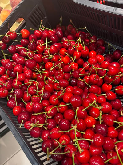 A black, slotted plastic crate is filled to the brim with numerous ripe red cherries, observed from a high-angle, close-up perspective. The cherries vary in color from bright red to a deeper, rich burgundy, and many retain their attached green stems, some individually and others in small clusters. The crate features a grid-like pattern of horizontal and vertical slits. In the upper left background, out of focus, several peach-colored fruits are partially visible within an adjacent container. A glimpse of a light-colored surface, possibly a floor or countertop, is discernible in the lower left of the frame. The scene presents a static display of fresh produce within a commercial environment in the city of Krustkalni, Latvia. No individuals are present.