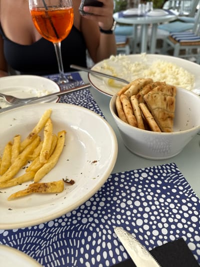 A high-angle, close-up shot captures a partially consumed meal at an outdoor restaurant in Mykonos, Greece. In the foreground, a white ceramic plate contains remnants of golden-brown french fries, lightly seasoned, with a few crumbs scattered on the plate. To its right, a white ceramic bowl holds multiple pieces of folded pita bread, sprinkled with green herbs. Beneath these items, a blue placemat with a white polka-dot and wave pattern is visible, with a silver serrated knife resting on a black napkin.
In the mid-ground, a person, likely female, is partially visible on the left, wearing a black top. Her left hand holds a dark smartphone, and a watch is visible on her right wrist. A tall stemmed glass containing an orange beverage (possibly an Aperol Spritz) with a dark straw is positioned in front of her. Further back, a white serving dish contains a large quantity of grated white cheese or a similar crumbly substance, with a lemon wedge and a silver serving utensil. Another partially cleared white plate with a fork lies between the foreground fries plate and the cheese dish. The background is softly blurred, showing other light-colored tables and chairs with blue and white striped cushions, indicative of a Mediterranean dining establishment.