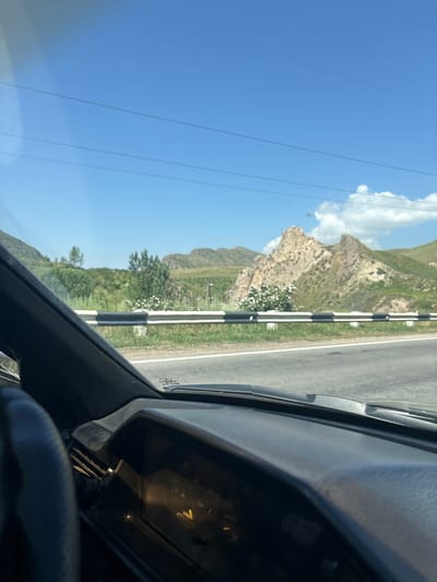 The image is a first-person perspective from inside a car, looking out through the windshield. The view encompasses a road, a guardrail, and a mountainous landscape. The sky is a clear blue with a few scattered clouds. In the foreground, the dashboard of the car is visible, including part of the steering wheel and the instrument cluster, which shows a yellow indicator light. The road is paved and appears to be a highway or main road. A black and white striped guardrail runs along the side of the road. Beyond the guardrail, there are green hills and mountains. One prominent rock formation in the distance has a jagged, pointed peak. Bushes and trees are scattered across the landscape. The overall scene suggests a scenic drive through a rural or mountainous area. Based on the prompt's instruction regarding Zangakatun, Armenia, this image depicts a view from a vehicle traveling on a road in or near Zangakatun, Armenia, with the characteristic mountainous terrain and clear blue sky of the region.