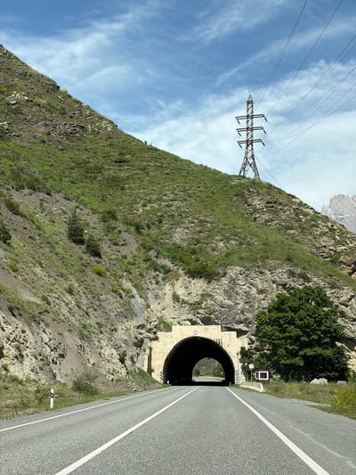The image depicts a highway leading into a tunnel carved through a mountainous landscape. The road is paved with asphalt and marked with white lines. A steep, grassy mountain slope rises to the left of the road, dotted with rocks and sparse vegetation. To the right, the tunnel entrance is visible, a large arched opening framed by concrete. Adjacent to the tunnel entrance, a dense green tree stands, and further to the right, a small, white booth with a dark square in its center is positioned. A tall power line tower stands atop the hill to the right of the tunnel, with its wires stretching across the sky. The sky is blue with scattered white clouds, and the overall environment suggests a rural or mountainous area. The location is identified as being in Mizur, Russia.