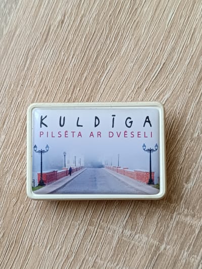 This is a rectangular magnet featuring a scenic photograph of a bridge. The magnet has a white border. The photograph depicts a street lined with red brick railings and old-fashioned lampposts. A lone figure, a person in a dark coat, is walking away from the viewer down the street. The scene is shrouded in a thick fog, suggesting a cool, misty atmosphere. The background is obscured by the fog, creating a somewhat mysterious or serene mood.

Above the image, the text "KULDĪGA" is printed in black capital letters. Below this, in smaller red capital letters, is the text "PILSETĀ AR DVĒSELI". The overall impression is a representation of Kuldīga, a town in Latvia, possibly highlighting its historic charm and tranquil ambiance, as suggested by the fog and the phrase "city with a soul". The time of day is ambiguous due to the fog, but the presence of lampposts might imply it's either early morning or late evening.