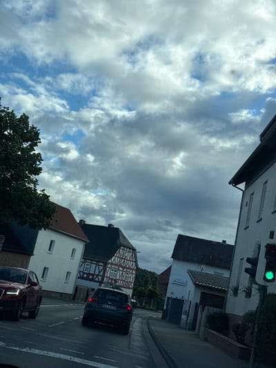 This is a photograph of a street scene in Volpertshausen, Germany. The image is oriented vertically, suggesting it was taken from inside a vehicle looking upwards and to the side.
The subjects include several buildings, a traffic light, and two cars. The buildings have traditional German architectural elements, with half-timbered facades visible on one structure. One building appears to be a business with signage that reads "Stromberg". The traffic light is green. The cars are a dark-colored hatchback and a red sedan. The red sedan's headlights are on, and the dark hatchback's taillights are illuminated.
The scene depicts a street with a paved road and a sidewalk. Trees are visible, with one large tree in the foreground. The sky is filled with dramatic clouds, suggesting cloudy or partly cloudy weather. The lighting indicates it is daytime, possibly late afternoon or early evening. The overall mood is neutral, capturing a typical street view.
There is visible text on the license plate of the dark hatchback, which appears to read "WEL-OG 88". The sign on the building reads "Stromberg". The traffic light has an illuminated green signal.