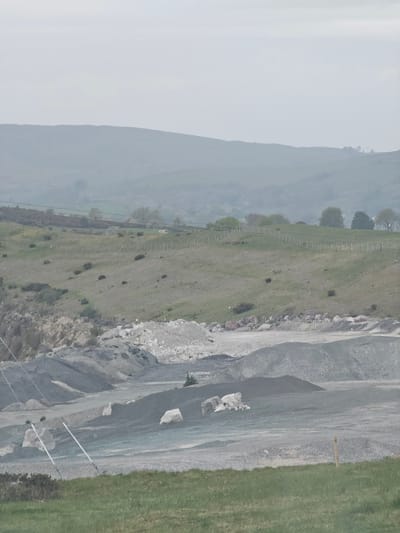 The image shows a view of a quarry located near Halkyn, United Kingdom. A partially visible mountain is shrouded in mist in the left background. A fence line separates a grassy field from the quarry, which is composed of grey rock and loose gravel. An exposed rock face is also visible. Power lines are visible.