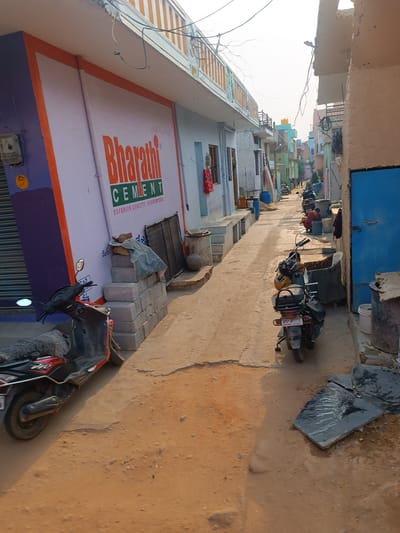 The image depicts a narrow street in Kothacheruvu, India. The street is unpaved and dusty, with buildings on either side. On the left, a building prominently features the "Bharathi CEMENT" logo. Parked nearby is a scooter. On the right side of the street, a blue door is visible. Further down the street, more buildings line the road, and several parked motorcycles can be seen. In the distance, there are glimpses of a water tank and other structures. The overall scene is characteristic of a residential area in an Indian town.