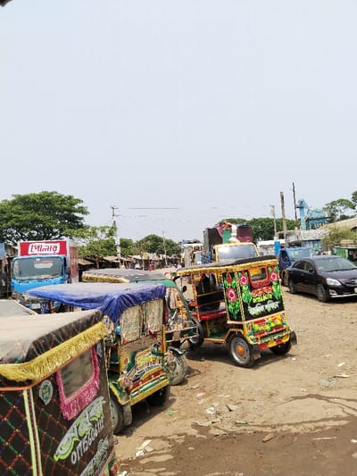 The scene depicts a bustling street in Pangasia, Bangladesh, captured from a slightly elevated perspective. The street is lined with colorful auto-rickshaws, known locally as "CNGs," parked along the side of the road. The auto-rickshaws are decorated with elaborate floral designs and adorned with fringes and tassels, showcasing the local culture and vibrant aesthetics of the area. Several other vehicles can be seen parked in the background, including a blue truck with a Bengali script advertisement on its side and a silver sedan.  The ground is covered in a layer of dirt and debris, which is common in the region. The photograph was taken in daylight under a clear sky, indicating a bright and sunny day.