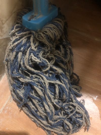 A close-up, high-angle shot shows a dirty, blue and beige string mop resting on a wooden floor. The mop head is densely packed with frayed strings, some appearing damp or matted with dirt. A portion of a blue plastic mop handle is visible at the top of the image, connecting to the mop head. The wooden floor is a light brown color with visible grain and appears slightly damp in areas, with some light reflecting off the surface. The background is dimly lit and appears to be a wall or another surface, also with a textured appearance. There are no people or animals present.