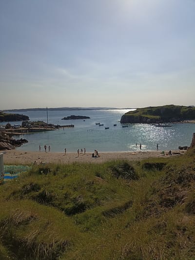 The image depicts a sunny day at a beach in Carnboy, Ireland. The scene is a wide shot of a sandy beach meeting the ocean, with a rocky outcropping on the left and a larger, grassy island in the distance on the right. In the foreground, tall, dry grass covers a slight incline.
On the beach, several people are visible. Closer to the water, a group of children and adults are either standing or walking. Further back on the sand, a few individuals are seated or interacting with each other. The water is clear and calm, with several small boats anchored in the bay. Some of these boats are near the shore, while others are further out towards the distant islands.
To the left of the image, there is a stone pier with a few people on it. Adjacent to the pier, there's a small building and some parked vehicles, suggesting a small harbor or port area. The sky is a clear, pale blue with a slight haziness near the horizon. The sun is bright, casting a shimmering reflection on the water's surface. The overall atmosphere is one of a relaxed summer day at the coast.