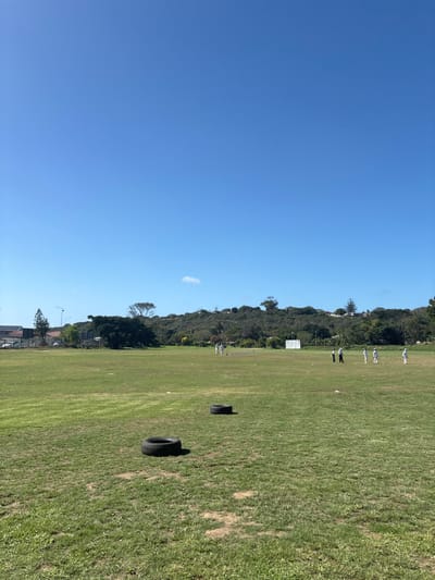 The image depicts an outdoor cricket ground under a clear, bright blue sky with one small cumulus cloud near the horizon. The location is within the Ndlambe Local Municipality, South Africa.

In the foreground and midground, a large, expansive green grass field occupies the majority of the frame, showing patches of dry or less dense grass. Two black vehicle tires are lying flat on the grass in the midground; one is closer to the viewer and slightly left of center, the other further back and to the right.

Approximately nine individuals are visible on the field in the background, primarily positioned in the central and right portions of the frame. They appear to be engaged in a cricket activity; most are wearing light-colored clothing, possibly cricket whites. A set of cricket wickets (stumps) is visible in the middle-left of the group. A large white cricket sight screen stands upright on the field further back, to the left of the main group of people.

The background features a prominent, green, tree-covered hill stretching across the horizon from the center to the right. Small residential structures or buildings are intermittently visible on the slopes and crest of this hill. To the far left background, parts of urban infrastructure are visible, including buildings with dark roofs, one featuring solar panels, and utility poles with streetlights. A dense cluster of trees and vegetation separates this urban area from the main cricket field.