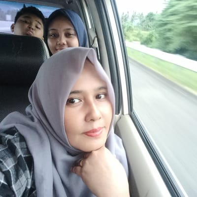 This image depicts a group of people traveling in a car in Cotkareung, Indonesia, captured during what appears to be a daytime journey. The main subject, positioned in the foreground, is a woman wearing a light grey hijab and a grey and black plaid shirt. She is looking directly at the camera with a calm, content expression and a slight smile, with her right hand gently resting under her chin, revealing a gold-colored ring on her finger.
Behind her, partially obscured by the car seat, are two other individuals. To the right, a young woman wearing a dark blue hijab is visible, also looking towards the camera with a faint smile. To the left of her, peeking from behind the headrest, a young boy is making a playful face, pouting his lips.
The scene is set inside a vehicle, indicated by the car seats and window frame. Through the car window, a blurred background suggests motion, indicating the car is traveling. Green foliage, likely trees and grass, lines what appears to be a road or highway, further emphasizing the sense of travel or a road trip. The lighting is natural and bright, suggesting it is daytime. The overall mood conveyed is one of relaxation and enjoyment during transit. There is no visible text in the image.