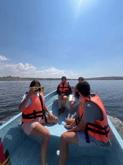 The image shows a group of people on a boat on a lake in the city of El Oasis Valsequillo, Mexico. The people are wearing life jackets and appear to be taking a boat trip. One person is holding a cell phone. The lake water is dark and rippled, and the sky is clear with a few clouds. A strip of land can be seen in the background.