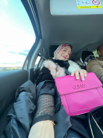 The image captures a vibrant moment inside a car, likely in Majmaa Tolba, Morocco, during daylight hours. A smiling woman is the primary subject, positioned in what appears to be a passenger seat, looking cheerfully towards the camera. She wears a patterned hijab in muted pinks and beiges, complementing a stylish coat featuring both black and light pink fur trim. Her right hand, adorned with rings, holds a striking fuchsia pink Guess handbag, clearly displaying the brand name "GUESS" embossed on its flap. Beneath her outer layers, a sheer, dark patterned sleeve, possibly part of a top or stocking, can be seen, intricately detailed with tiny heart shapes.

In the upper right background, another person is partially visible, wearing a brown coat and a dark hat with fur, suggesting fellow passengers. The car's interior is evident, with a sun visor in the upper left corner prominently displaying an "AIRBAG" warning label and safety symbols. Through the car window on the left, a bright, clear sky dominates, with glimpses of a distant, undulating landscape, indicative of an outdoor setting. The scene exudes a sense of pleasant travel and happiness.