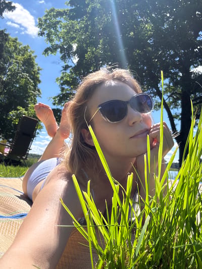 In Lendži, Latvia, a woman is lying on a mat outdoors on a sunny day, with her body angled towards the camera and her face in profile. She is wearing sunglasses and has her chin resting on her hand. Tall grass is in the foreground, partially obscuring her. In the background, there are trees and a bright blue sky with some clouds. A lounge chair is visible to the left, and a body of water can be seen in the distance. The woman's feet, wearing striped bikini bottoms, are also visible.