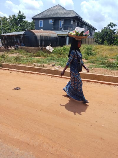 A woman walks along a dirt road in Iseyin, Nigeria, carrying a tray of fruit on her head. She is wearing a long, patterned blue dress and a headscarf, and a black bag is slung over her shoulder. The fruit on the tray appears to include watermelon slices, cucumbers, and possibly other fruits. The sun is shining brightly, casting a long shadow of the woman onto the road, suggesting it is likely midday.

In the background, a large, grey house with a dark tiled roof stands prominently. The house has multiple windows and a balcony. To the left of the house is a long, corrugated metal structure with a fence in front of it. Trees and green vegetation are visible on either side of the road and behind the structures. The road itself is composed of orange-brown dirt with some scattered stones and debris. A concrete curb runs along the edge of the road. On the road, to the left of the woman, a pair of dark sandals lies discarded. The sky is partly cloudy with a bright sun. The overall scene depicts a typical day in a Nigerian town, with a vendor going about her business under the open sky.