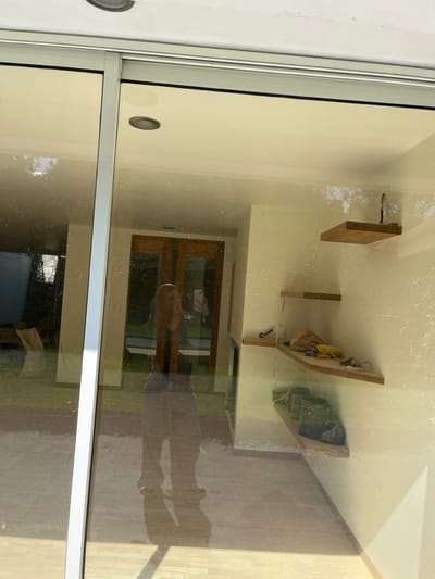 The image shows the reflection in a glass door or window. Inside, there are wooden shelves on the wall, a wooden door, and possibly a couch or other furniture. Outside, a person's shadow is cast on a light-colored wooden floor or surface. The reflection shows a distorted view of both the interior and exterior spaces.