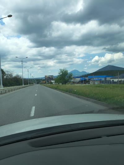The image depicts a view from a car driving down a two-lane asphalt road in Goryachevodsky, Russia, on a cloudy day. The road has white dashed lines marking the lanes. On the left side of the road, there is a metal guardrail, followed by trees and vegetation. On the right side, there is a ditch with tall grass, a sidewalk, and then buildings and houses with blue roofs, interspersed with green trees. In the distance, there are several mountains, one of which has a tower on its summit. A billboard advertising cars is visible on the right side of the road. Streetlights line the left side of the road. The sky is filled with large, grey clouds with patches of blue sky visible. The car's dashboard is partially visible in the foreground.