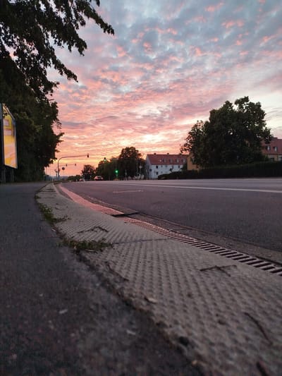 The image captures a low-angle view of a street corner in Kitzingen, Germany, during a sunset. The sky is filled with dramatic clouds painted in shades of pink, orange, and grey, indicating the end of the day. In the foreground, a grey asphalt road curves to the left, bordered by a textured metal grate along the sidewalk. Green traffic lights are visible in the distance, suggesting an intersection. Trees with dark green foliage line the street on the left and right, and residential buildings with red-tiled roofs are present on the right side of the road. The overall scene is one of urban life transitioning into twilight, with the natural beauty of the sunset contrasting with the man-made environment of the city.