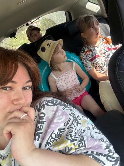 The image is a selfie taken inside a car, likely in the city of Аряж, Russia, with the sun casting light through the rear window. Three children and a woman are visible. In the foreground, a woman with reddish-brown hair is looking at the camera, her hand near her mouth. Behind her, a young girl wearing a straw hat with a cartoon face is seated, and next to her, another girl in a white t-shirt with graffiti-like print. Further back, a boy wearing a baseball cap is looking upwards. The car's interior is dark, with black seats and a partially visible sunshade. Outdoor scenery, including greenery and buildings, is visible through the rear window.