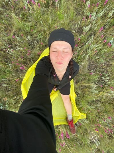 A high-angle, waist-up selfie shows a person with closed eyes, wearing a black beanie, black long-sleeved shirt, and black shorts, lying on a bright yellow ground cover in a grassy field with scattered purple flowers. Their right arm is extended, holding the camera. A person in black pants is partially visible in the lower left corner, suggesting they are taking the photo. The location is identified as Ziyanchurino, Russia.