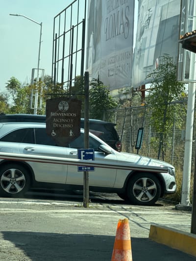 A white car parks adjacent to a pedestrian sign indicating "Ascenso y Descenso." The car, partially obscured, showcases its wheels and side profile. Behind, there's a large promotional banner alongside a metal fence. The ground is paved, featuring a traffic cone near the car. The sky appears clear, and trees are scattered in the background, providing a glimpse of greenery amidst the urban environment.
