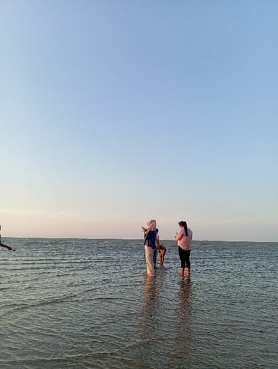 The image depicts a scene at a shallow body of water in the city of Tritunggal, Indonesia. The sky is clear, light blue, and fades to a lighter tone near the horizon, occupying approximately two-thirds of the frame. The lower third consists of calm, rippled water, revealing an undulating sandy or muddy bottom.
Four individuals are present in the water. On the far left, a portion of a person's left arm and shoulder is visible. In the center, a person wearing a light-colored garment, a blue outer layer, and a hijab stands, holding a mobile device horizontally, aimed towards the right. To their immediate right, another individual, partially obscured and dressed in dark clothing, is bent over, facing away from the camera, with their lower body submerged or obscured. On the far right, a fourth person, wearing a light pink long-sleeved shirt and dark pants, stands with their back to the viewer, facing the central figures. The water level reaches approximately the mid-calf of the standing individuals. The horizon line is flat and clear.