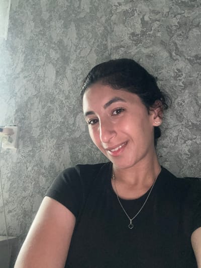 A young woman with dark hair pulled back and a black shirt is looking at the camera. She is wearing a silver necklace with a clover-shaped pendant and a small stud earring with three small stones. The background is a textured gray wall with a light shining from the left side of the frame, illuminating a white electrical outlet. The woman is smiling, and her right arm is visible, resting near her chest. The image appears to have been taken indoors. The location is identified as Dondukovskaya, Russia.