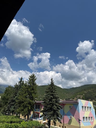 The image presents a bright, sunny daytime scene in a mountainous region, consistent with Rila, Bulgaria. The upper portion of the frame is dominated by a clear blue sky, interspersed with numerous large, fluffy white cumulus clouds.

In the background, a large mountain or hill rises, densely covered in dark green foliage, suggesting a forest. Several tall, dark green coniferous trees, likely pines or spruces, are prominent in the mid-ground, partially obscuring the view of the buildings.

On the right side of the frame, a distinctive multi-story building stands out with its facade painted in a vibrant diamond pattern of pastel colors, including pink, light blue, and yellow. The building features several rectangular windows with white frames, and two air conditioning units are visible on its exterior walls. A glimpse of another building with a dark brown roof is seen behind the colorful structure. The foreground includes various green bushes and foliage.

There are no people visible, and no specific activities are taking place; the scene captures a static landscape and architectural view. The bright sunlight and clear sky indicate it is likely late morning or early afternoon. No visible text is present in the image. A dark, possibly architectural element, frames the bottom left corner of the sky, suggesting the photo was taken from a vantage point that includes part of a structure in the immediate foreground.
