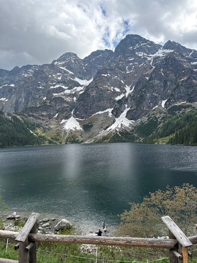 The image captures a mountain lake scene. A large, craggy mountain with snow patches dominates the left side of the frame, under a cloudy sky. The right side features a dark, still lake reflecting light. A wooden railing is visible in the foreground, partially obscuring the shoreline. People are on the edge of the lake. This scene could be in the city of Brzegi, Poland.
