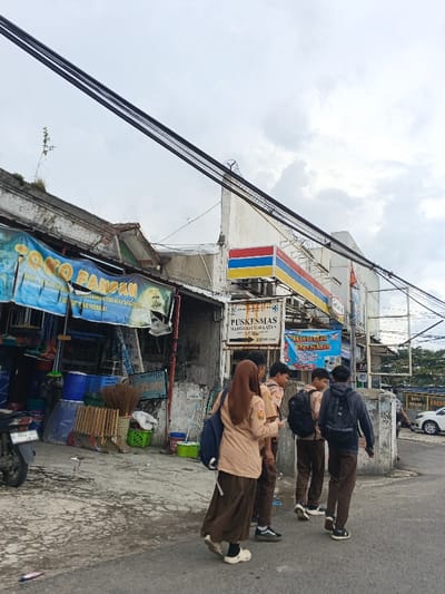 A street scene depicts a group of four students walking on a cracked pavement. They are wearing school uniforms, and each has a backpack. Behind them are buildings, one with a worn blue banner and another with a red, yellow, and blue storefront sign, likely a convenience store. Power lines crisscross the overcast sky. A motorcycle is parked beside the first building, and a car is visible in the distance.