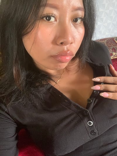 A close-up, high-angle, selfie-style, medium shot shows a person with dark hair, fair skin, and full lips, wearing a black, collared shirt with buttons down the placket. The person has long, dark hair that falls loosely around their face and shoulders. Their eyes are dark and looking towards the camera. Their lips are glossy and appear to be slightly parted. A thin gold chain necklace is visible around their neck. The person's right hand, with manicured nails featuring a pink base with black polka dots, is resting on their chest, near the buttons of the shirt. The background is out of focus, showing a patterned wallpaper and a hint of a textured fabric in red and gold. There is no discernible information about the location in Jajag, Indonesia.