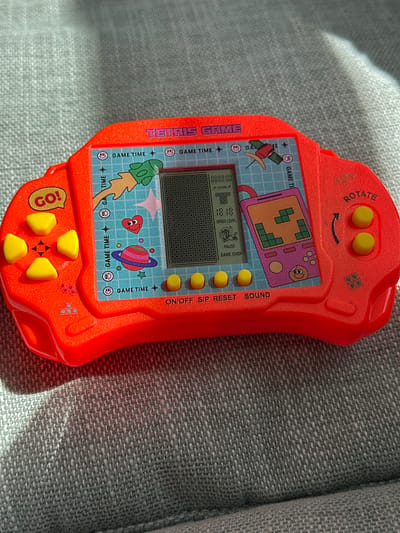 A red, handheld electronic game, likely a Tetris clone, rests on a grey, woven fabric surface. The game console features a screen displaying a Tetris game in progress, along with game time, score, speed level, pause, and game over indicators. It has a directional pad on the left with a "GO!" button and two yellow action buttons on the right labeled "ROTATE." The console is adorned with cartoonish graphics including rockets, planets, and smiley faces. The fabric surface appears to be upholstery, possibly from a sofa or chair. The lighting suggests it is daytime, with sunlight casting shadows. There is no indication of the specific location in Moortown, Ireland; the scene is isolated to the object and its immediate surroundings.