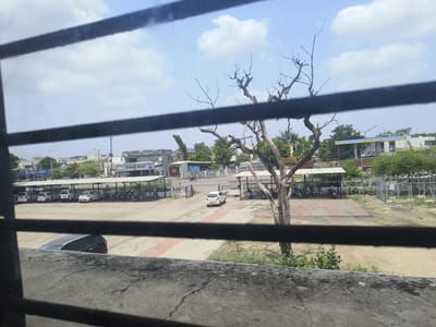 A medium-wide shot captures an outdoor scene through a barred window. In the foreground, a concrete ledge with visible cracks runs horizontally across the bottom. Behind it, a dirt lot is partially visible. A dark-colored SUV is parked at an angle in the dirt lot, with its rear end facing the viewer.
Beyond the dirt lot, a paved area features a covered parking structure with white roofs supported by poles. Several white cars and motorcycles are parked under this shelter. To the left of the parking structure, a building with "HDFC BANK" signage is visible. Further back, a gas station with a blue and yellow canopy is prominent.
A leafless tree with gnarled branches stands in the middle ground, to the right of the covered parking. In the background, a street with a few buildings and trees lines the horizon. The sky above is blue with scattered white clouds. The scene appears to be located in Dhandhuka, India, based on the architectural style and signage. No people are clearly identifiable in the image.