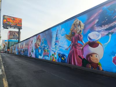 Here is a detailed description of the image:
The image shows a long mural painted on a wall in the city of La Victoria, Peru. The mural features characters and themes from the Super Mario Galaxy video game series. Princess Peach and Toad are depicted, with other characters and scenes from the game also visible along the length of the wall.
The mural is painted on a wall that runs along a street, which occupies the left half of the frame. There are cars and a person riding a motorbike far in the distance on the street. Billboards and buildings are visible in the background. One billboard displays a Coca-Cola advertisement, and another features "Fuente Alta De Fibra".