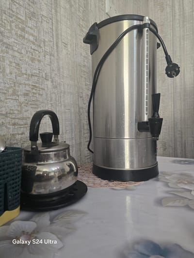 This is a close-up, horizontally oriented photograph showcasing kitchen appliances on a patterned tablecloth and a wooden-textured surface. The primary subjects are a large, silver water dispenser with a black spout and a smaller, shiny silver kettle with a black handle.
The water dispenser is positioned upright, taking up the left side of the frame. Its metallic body is cylindrical, with a black rim and base. A black power cord with a European-style plug is visible, extending from the top of the dispenser and curving downwards. A translucent water level indicator with markings is integrated into the side of the dispenser.
Below and to the right of the water dispenser sits the silver kettle. It has a rounded body, a black lid with a knob, and a prominent black handle attached by metal brackets. The kettle rests on a black coaster, which is itself placed on a white and grey patterned tablecloth with large floral designs in pale colors. A portion of another dark green object with yellow accents is visible in the bottom right corner, possibly part of a kitchen utensil or appliance.
The background consists of a light-colored, wood-grain patterned surface on the left and the floral tablecloth on the right. The lighting suggests an indoor setting, possibly during the daytime. No people are visible, and there are no strong indicators of the time of day or weather conditions.
A visible text watermark in the lower right corner reads "Galaxy S24 Ultra". The image does not appear to depict a specific event or activity, but rather a static arrangement of kitchen items. The overall impression is one of everyday domesticity.