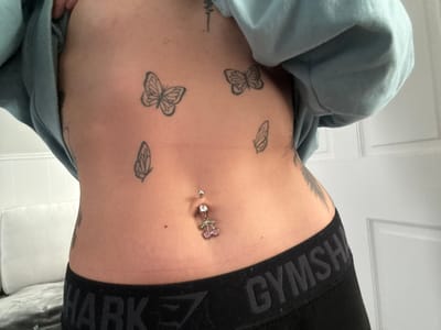 A close-up studio shot captures a person's torso and midriff, revealing skin decorated with several small black butterfly tattoos and what appears to be a black feather tattoo on the side. A belly button piercing with a cherry charm and a dangling gem is prominently visible. The person is wearing a black athletic band with the word "GYMSHARK" repeatedly printed on it. A light blue hoodie is draped around the shoulders and upper chest. The background features a white paneled door and a white cushion.