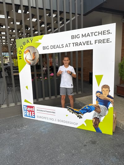 In the city of Kulata, Bulgaria, a young boy is posing for a photo in a large, white, rectangular photo booth frame set up outdoors. The frame has green and yellow accents and features text that reads "HOORAY BIG MATCHES. BIG DEALS AT TRAVEL FREE." and "EUROPE'S NO.1 BORDERSHOP". A soccer ball is depicted on the left side of the frame, and an illustration of a soccer player in a blue and white uniform is on the right. The boy is wearing a white t-shirt, grey shorts, and soccer cleats. He is smiling and pointing at himself with both hands. In the background, there are outdoor seating areas with tables and chairs, and some potted plants. The ground is paved with a dark, textured material.