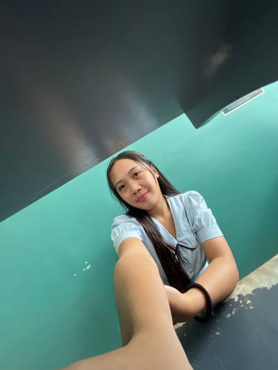A young woman with long dark hair and light skin is taking a selfie. She is wearing a light blue collared shirt with short puffed sleeves. She has a black bracelet on her left wrist. She is leaning on a dark gray surface, with a teal wall behind her. A black ceiling or overhang is above her and partially obscures the view. There are some small white splatters on the teal wall. A sign with text is visible on the teal wall. The image was taken indoors.