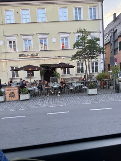 The image depicts an outdoor seating area of a pizzeria, "MARTHA PIZZAREI," located in Fürstenfeldbruck, Germany. Several people are seated at tables covered with green and white checkered tablecloths, appearing to be dining or socializing. The building behind the seating area is a light yellow facade with multiple white-framed windows. A tree grows in front of the building, extending branches over the outdoor seating. Umbrellas provide shade over some of the tables. The ground is paved with cobblestones next to the building, and a paved road with white lane markings is visible on the right side of the image. String lights are visible, suggesting it might be late afternoon or evening, as they are illuminated. The overall atmosphere appears relaxed and casual. The text "MARTHA PIZZAREI" is prominently displayed on the building facade and on a wooden crate in the foreground.